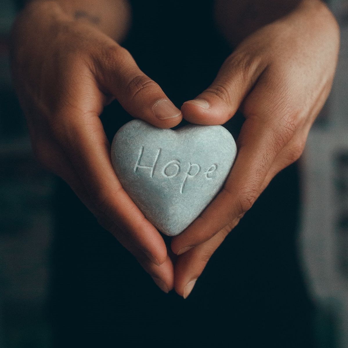 Hands clasp around a light gray, heart-shaped rock the has "Hope" engraved into 
