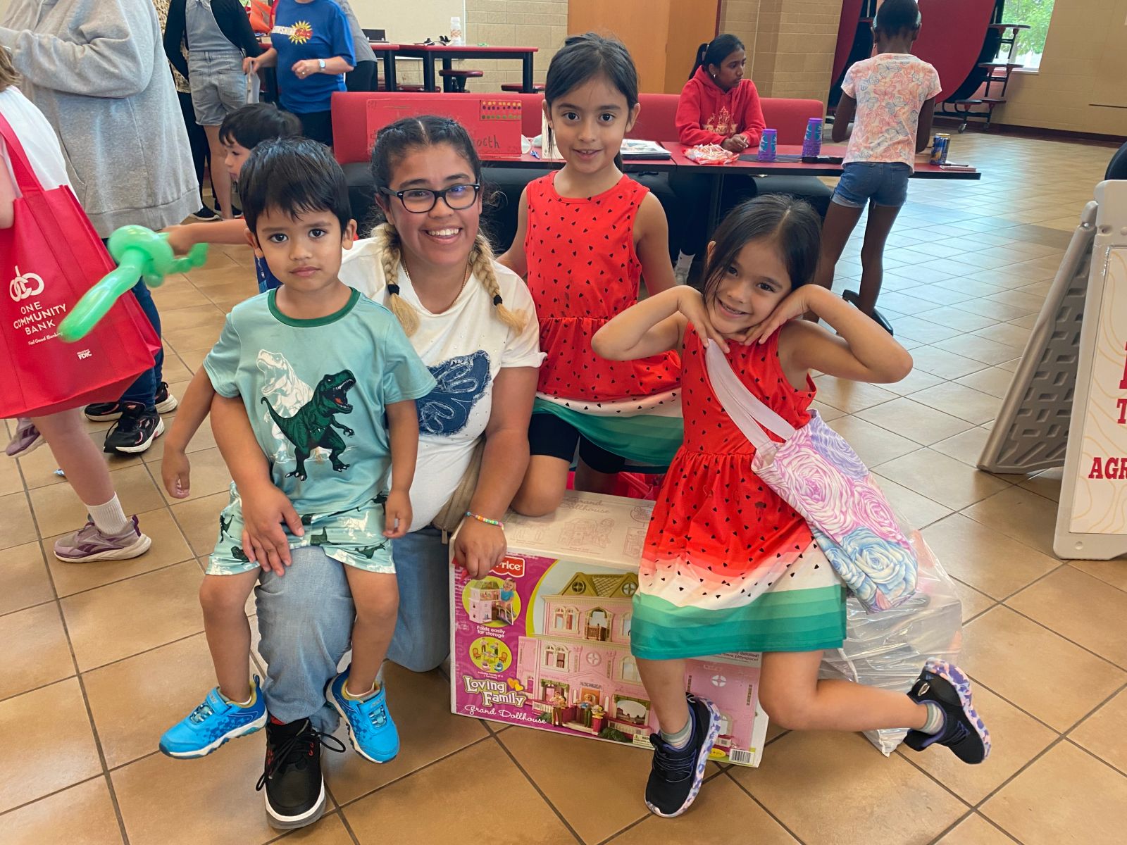 A woman kneels on the floor by a box with a picture of a dollhouse. A little boy in a dinosaur shirt sits on her knee, and two little girls in matching watermelon dresses sit on the box next to her.