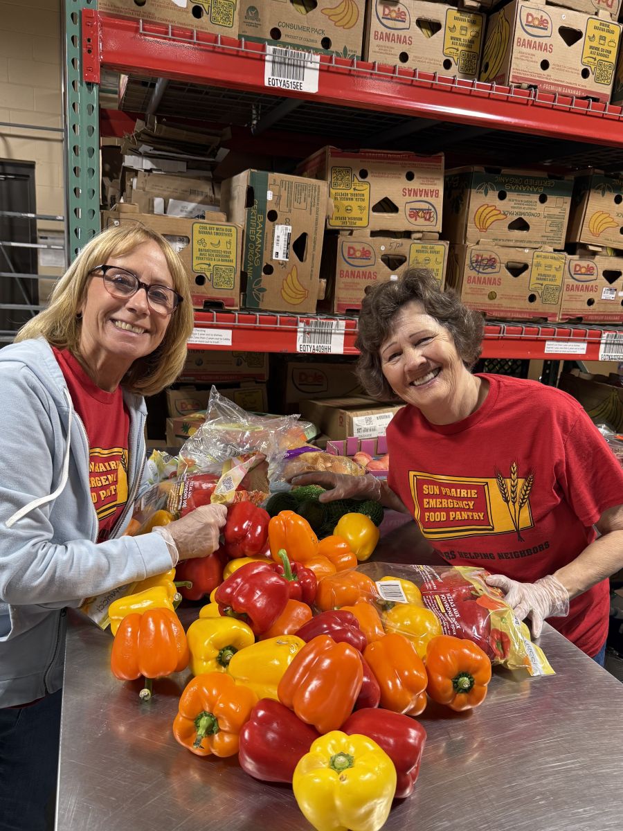 Two women wearing red Sun Prairie Food Pantry shirts smile beside a stainless steel table with a pile of red, orange, and yellow bell peppers. 