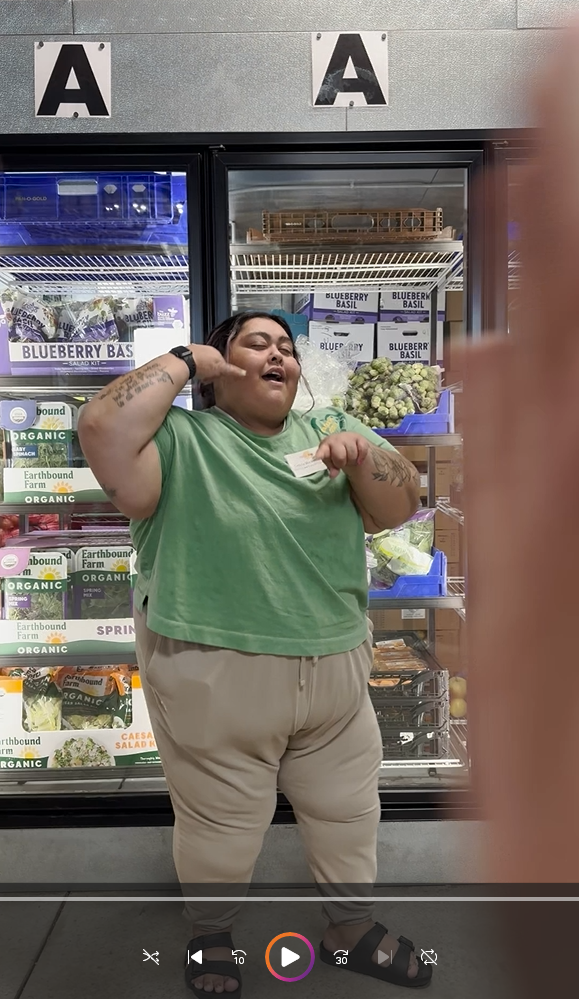 A woman in a green shirt and tan pants stands in front of a food pantry cooler full of food, ready to dance. There is a play button indicating this is linked to a video. 